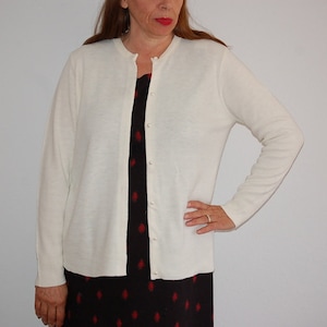 以下が含まれることがあります： ボタン留めのフロントを持つ、白い長袖カーディガン。カーディガンは柔らかく、ニット素材で作られています。