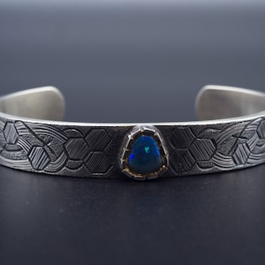May include: Silver cuff bracelet with a geometric pattern and a triangular, iridescent blue opal gemstone set in a gold bezel. The bracelet has an open design and is displayed against a dark background.