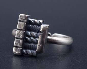 May include: A silver ring with a textured, geometric design. The ring is made of metal and has a unique, modern style.