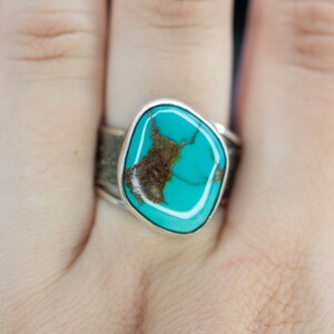 Op de afbeelding: Een zilveren ring met een turquoise steen in een vierkante vorm. De steen heeft een bruine matrix en een blauwgroene kleur.