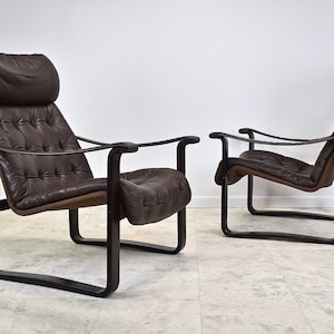 May include: Two brown leather lounge chairs with a dark wood frame. The chairs have a tufted design and are angled slightly towards each other.