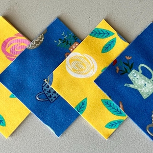 May include: Four square fabric coasters with floral and teapot patterns. Two coasters are yellow with blue and pink floral designs. The other two are blue with white and yellow teapot designs. Green leaves accent the designs.