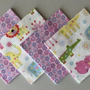 May include: A collection of five square fabric napkins. The napkins feature a variety of patterns, including floral designs and cartoon animals such as lions, elephants, and giraffes. The color palette includes pink, yellow, and blue.