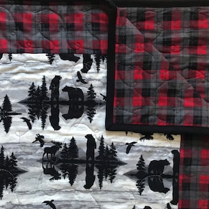 May include: A quilted throw blanket with a black, red, and grey plaid pattern on one side and a white and grey pattern with black silhouettes of trees, bears, and moose on the other side.