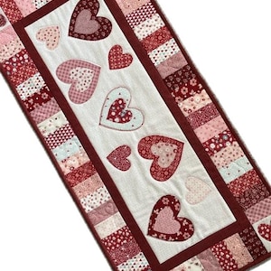 Może przedstawiać: Prostokątny, pikowany bieżnik na stół z białym środkowym panelem ozdobionym aplikowanymi sercami o różnych rozmiarach i wzorach. Obramowanie składa się z kwadratów patchworkowych w odcieniach czerwieni, różu i bieli. Przedmiot dekoracyjny.