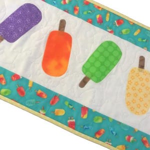 Może przedstawiać: Biały pikowany bieżnik stołowy z turkusowym obramowaniem, na którym widnieje powtarzający się wzór kolorowych lodów. Bieżnik ozdobiony jest czterema naszytymi lodami w kolorze fioletowym, pomarańczowym, zielonym i żółtym.