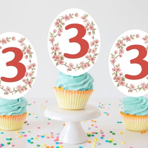 May include: Three cupcakes with blue frosting and yellow liners. Each cupcake has a white topper with a red number 3 surrounded by a floral wreath. Colorful sprinkles are scattered around the cupcakes, which are on a white surface.