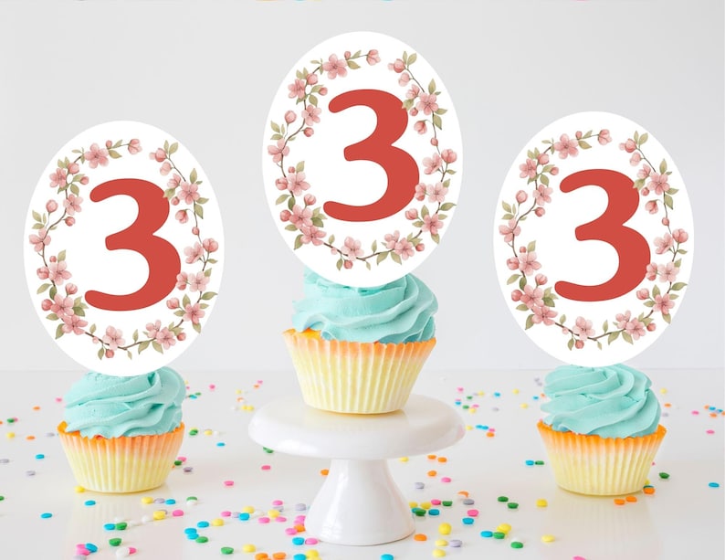 May include: Three cupcakes with blue frosting and yellow liners. Each cupcake has a white topper with a red number 3 surrounded by a floral wreath. Colorful sprinkles are scattered around the cupcakes, which are on a white surface.