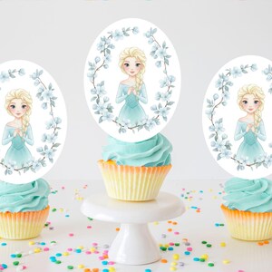 Op de afbeelding: Drie cupcakes met blauwe glazuur en gele vormpjes zijn bekroond met ovale afbeeldingen van een blonde vrouw in een blauwe jurk, omringd door blauwe bloemen. E&eacute;n cupcake staat op een witte taartstandaard, met hagelslag verspreid.