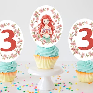 May include: Three cupcakes with blue frosting and yellow liners, topped with decorative toppers. Two toppers feature the number 3 in red, surrounded by pink floral wreaths. The center cupcake has a topper with a cartoon mermaid and floral accents.