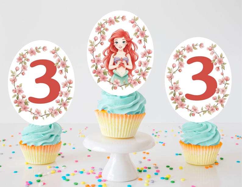 May include: Three cupcakes with blue frosting and yellow liners, topped with decorative toppers. Two toppers feature the number 3 in red, surrounded by pink floral wreaths. The center cupcake has a topper with a cartoon mermaid and floral accents.