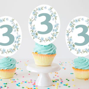 Op de afbeelding: Drie cupcakes met blauwe glazuur en gele vormpjes zijn bekroond met ovale decoraties met het nummer 3 in lichtblauw, omgeven door een bloemenkrans. Kleurrijke hagelslag is verspreid rond de cupcakes.