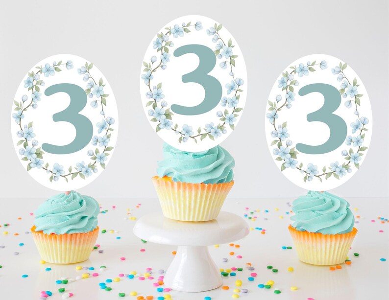 Op de afbeelding: Drie cupcakes met blauwe glazuur en gele vormpjes zijn bekroond met ovale decoraties met het nummer 3 in lichtblauw, omgeven door een bloemenkrans. Kleurrijke hagelslag is verspreid rond de cupcakes.