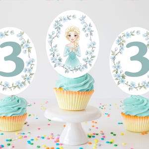 Op de afbeelding: Drie cupcakes met blauwe glazuur en gele vormpjes zijn versierd met toppers. Twee toppers hebben het blauwe nummer 3, omringd door blauwe bloemen. De middelste cupcake heeft een topper met een cartoonafbeelding van een blond meisje in een blauwe jurk, omringd door blauwe bloemen. Kleurrijke hagelslag is verspreid.