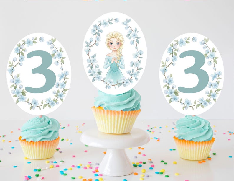 Op de afbeelding: Drie cupcakes met blauwe glazuur en gele vormpjes zijn versierd met toppers. Twee toppers hebben het blauwe nummer 3, omringd door blauwe bloemen. De middelste cupcake heeft een topper met een cartoonafbeelding van een blond meisje in een blauwe jurk, omringd door blauwe bloemen. Kleurrijke hagelslag is verspreid.