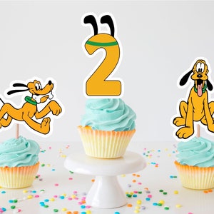 Puede incluir: Tres cupcakes decorados con un número dos amarillo, un perro de dibujos animados amarillo y naranja corriendo y un perro de dibujos animados amarillo y naranja sentado. Los cupcakes están cubiertos con glaseado azul claro y tienen forros de cupcakes amarillos.