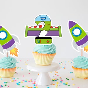 Op de afbeelding: Drie cupcakes versierd met groene, paarse en grijze rakettoppers. De middelste cupcake heeft een groene, paarse en rode topper met het cijfer "2" en de tekst "LIGHTYEAR" erop.