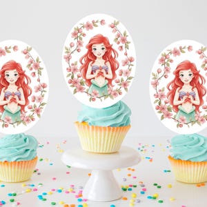 May include: Three cupcakes with light blue frosting and yellow liners are decorated with oval toppers featuring a red-haired cartoon character surrounded by pink flowers. The cupcakes sit on a white surface with colorful sprinkles.