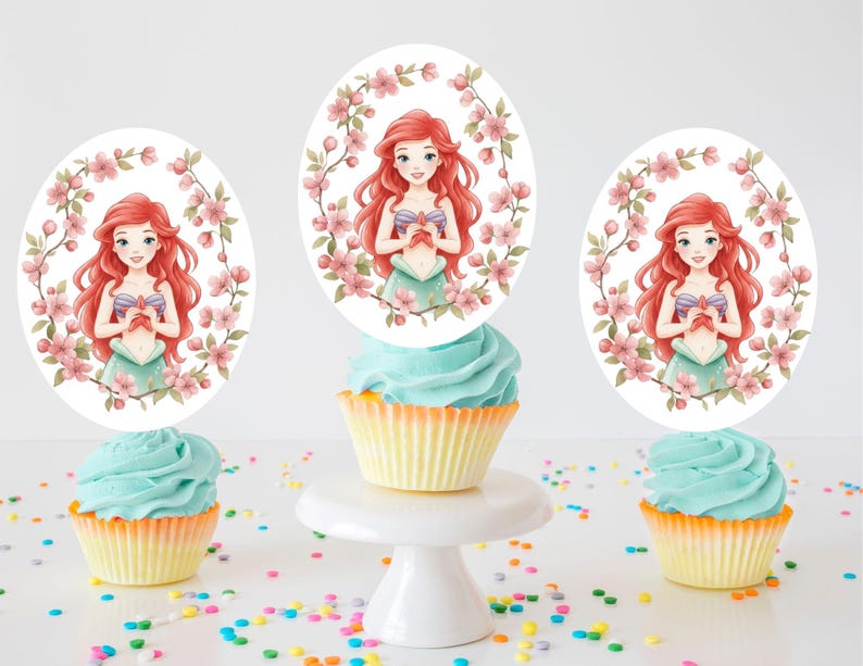 May include: Three cupcakes with light blue frosting and yellow liners are decorated with oval toppers featuring a red-haired cartoon character surrounded by pink flowers. The cupcakes sit on a white surface with colorful sprinkles.