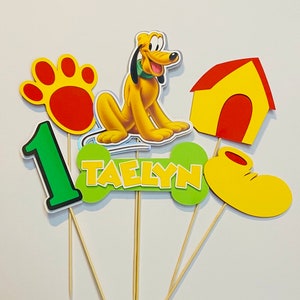 Pode incluir: Um conjunto de seis toppers para bolo com um cão de desenho animado, um osso, uma pegada de pata, uma casinha de cachorro, um sapato e o número um. O osso tem o nome "Taelyn" escrito nele. Os toppers são amarelos, verdes, vermelhos e laranja.