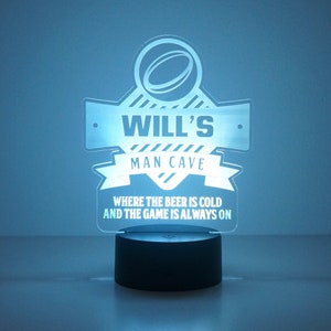 May include: A blue illuminated sign with the text "WILL'S MAN CAVE WHERE THE BEER IS COLD AND THE GAME IS ALWAYS ON". The sign features a stylized image of a football.