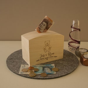 May include: A wooden money box with a teddy bear design. The box has a slot for coins and notes. The text "Jess & Ryan New Baby Fund" is engraved on the front of the box.