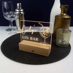 May include: A wooden sign with a clear acrylic panel that lights up. The panel has a gold outline of a gin and tonic cocktail and the text "Carly's Gin Bar".