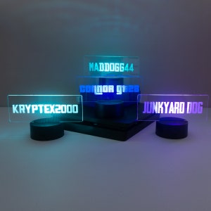 May include: Four acrylic signs with different names on them, lit up in different colours. The signs are on black bases and are arranged in a pyramid shape. The names on the signs are "MADDGG44", "connor gta28", "KRYPTEX2000", and "JUNKYARD DOG".