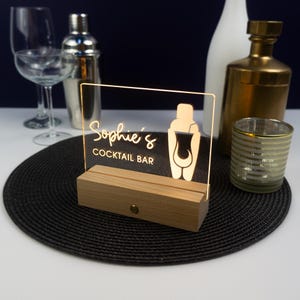 Könnte beinhalten: Ein Holzschild mit einem transparenten Acryleinsatz, der leuchtet. Das Schild trägt die Aufschrift "Sophie's Cocktail Bar" und zeigt eine Silhouette eines Cocktailshakers. Das Schild steht auf einem schwarzen runden Untersetzer.