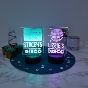May include: Two personalized LED light signs with a retro disco theme. The sign on the left features a boombox and the text "Stacey's Kitchen Disco". The sign on the right features a disco ball and the text "Lizzie's Kitchen Disco".