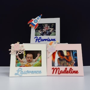 May include: Three personalised photo frames. One white frame has a rocket ship, the name "Harrison" in blue, and a photo. Another white frame has an octopus, the name "Lawrence" in blue, and a photo. A pink frame has a fairy, the name "Madeline" in red, and a photo.