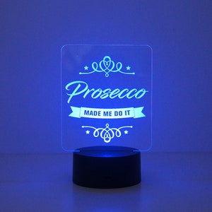 May include: A clear acrylic sign with the text "Prosecco Made Me Do It" in a decorative font. The sign is illuminated from behind with a blue light and sits on a black base.
