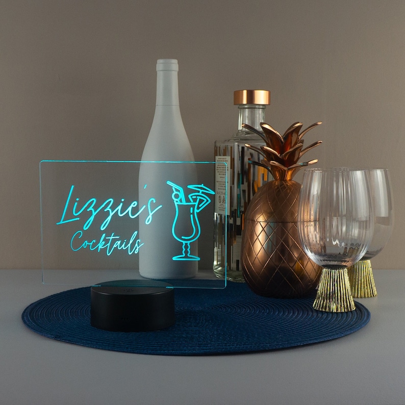 May include: A teal blue neon sign with the text "Lizzie's Cocktails" and a cocktail glass icon. The sign is on a black base and is sitting on a blue placemat. There are two clear glasses and a pineapple-shaped container on the table.