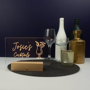 May include: A clear acrylic sign with a wooden base, illuminated with warm white LED lights. The sign reads "Josie's Cocktails" in gold script with a cocktail glass icon.