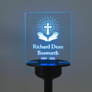 Peut inclure: Lampe commémorative solaire avec un panneau carré transparent. Le panneau affiche une croix, des mains et le texte "Richard Dean Bosworth 1950-2001" en blanc. La lampe a une base et une tige noires.