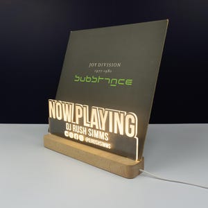 May include: A lit "Now Playing" record display stand. The stand has a wooden base and a clear acrylic front with the words "NOW PLAYING" in white. The display holds a Joy Division album cover. The album cover is dark green with white and green text.