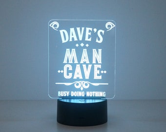 May include: A rectangular, illuminated sign with the text "DAVE'S MAN CAVE" in white, decorative lettering. Below the text is "BUSY DOING NOTHING". The sign is mounted on a black base, emitting a soft, blue glow.