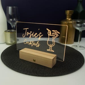 May include: A clear acrylic sign with a wooden base, illuminated from behind. The sign reads "Josie's Cocktails" with a stylized cocktail glass graphic.