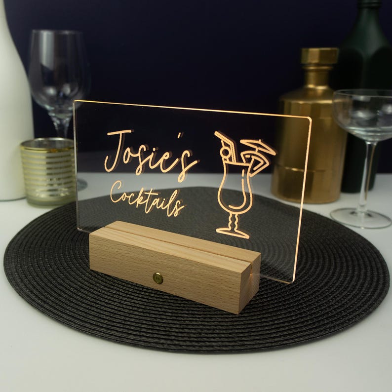 May include: A clear acrylic sign with a wooden base, illuminated from behind. The sign reads "Josie's Cocktails" with a stylized cocktail glass graphic.