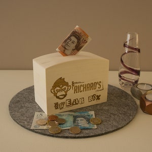May include: A wooden swear box with a monkey graphic and the text "Richard's Swear Box". The box has a slot in the top and is filled with coins and a British ten pound note.