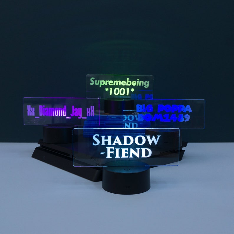 May include: A black gaming console with four different coloured acrylic signs on top. The signs have the text "Supremebeing *1001*" in green, "Xx_Diamond_Jay_xX" in purple, "BIG POPPA DOWNSMASH89" in blue, and "SHADOW -FIEND" in blue.