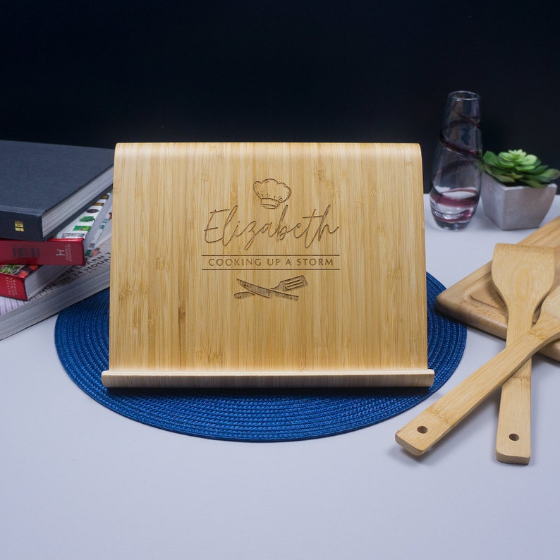 K&ouml;nnte beinhalten: Ein personalisierter Bambus-Kochbuchst&auml;nder mit dem Namen "Elizabeth" und dem Text "Cooking Up A Storm". Der St&auml;nder ist mit einer Kochm&uuml;tze und gekreuzten K&uuml;chenutensilien graviert.
