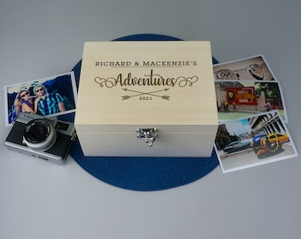May include: A wooden box with a hinged lid and a silver latch. The box is engraved with the text "Richard & Mackenzie's Adventures 2021" and has a decorative arrow design.