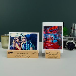 May include: Two photo displays with wooden bases. One holds a photo of a couple with the text "THE ADVENTURES OF Josh & Izzy" and a mountain and airplane graphic. The other displays a photo of a night market with the text "HONG KONG 2022 Temple Street Night Market".
