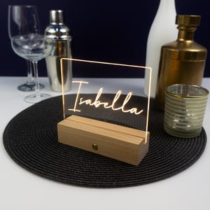 Peut inclure: Une plaque en acrylique transparente avec le nom "Isabella" illuminé d'une lumière blanche chaude. La plaque est sur un socle en bois et repose sur un set de table noir.