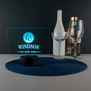 May include: A blue illuminated sign with the text "Windsor" and the words "Cocktails - Games - Sunday Roast" below. The sign is on a black base and is sitting on a blue placemat.