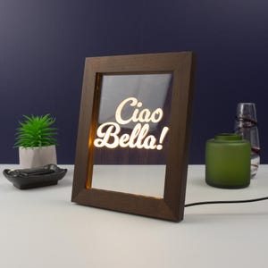 Pode incluir: Uma moldura de madeira marrom com uma frente de vidro transparente. A moldura é iluminada por trás, iluminando o texto branco "Ciao Bella!" no interior.
