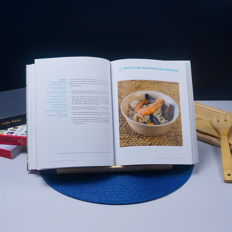K&ouml;nnte beinhalten: Offenes Kochbuch mit einem Rezept f&uuml;r Reisnudeln und Meeresfr&uuml;chte. Die Seite zeigt eine Nahaufnahme des Gerichts mit Reisnudeln, Garnelen, Muscheln und anderen Meeresfr&uuml;chten in einer wei&szlig;en Sch&uuml;ssel. Der Rezepttitel lautet "Reisnudeln und Meeresfr&uuml;chte" und die Zubereitungszeit betr&auml;gt 5 Minuten und die Garzeit 10 Minuten.