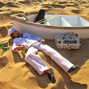 Puede incluir: Un barco de juguete blanco con una cubierta de madera y una barandilla de cuerda se encuentra en una playa de arena. Una muñeca de marinero de juguete con un uniforme blanco y un sombrero de marinero yace en la arena junto al barco. Dos botellas de vidrio verde están en la arena cerca de la muñeca.