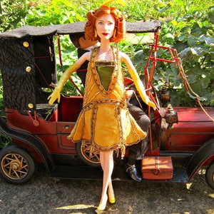 Puede incluir: Una muñeca con un vestido amarillo y verde con detalles dorados, sentada en un coche antiguo. La muñeca tiene el pelo rojo y lleva zapatos amarillos.